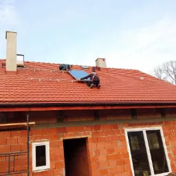 Ekipa dekarzy montuje panele słoneczne na dachu pokrytym czerwoną dachówką, widoczny budynek w trakcie budowy z cegły oraz fragment rusztowania.