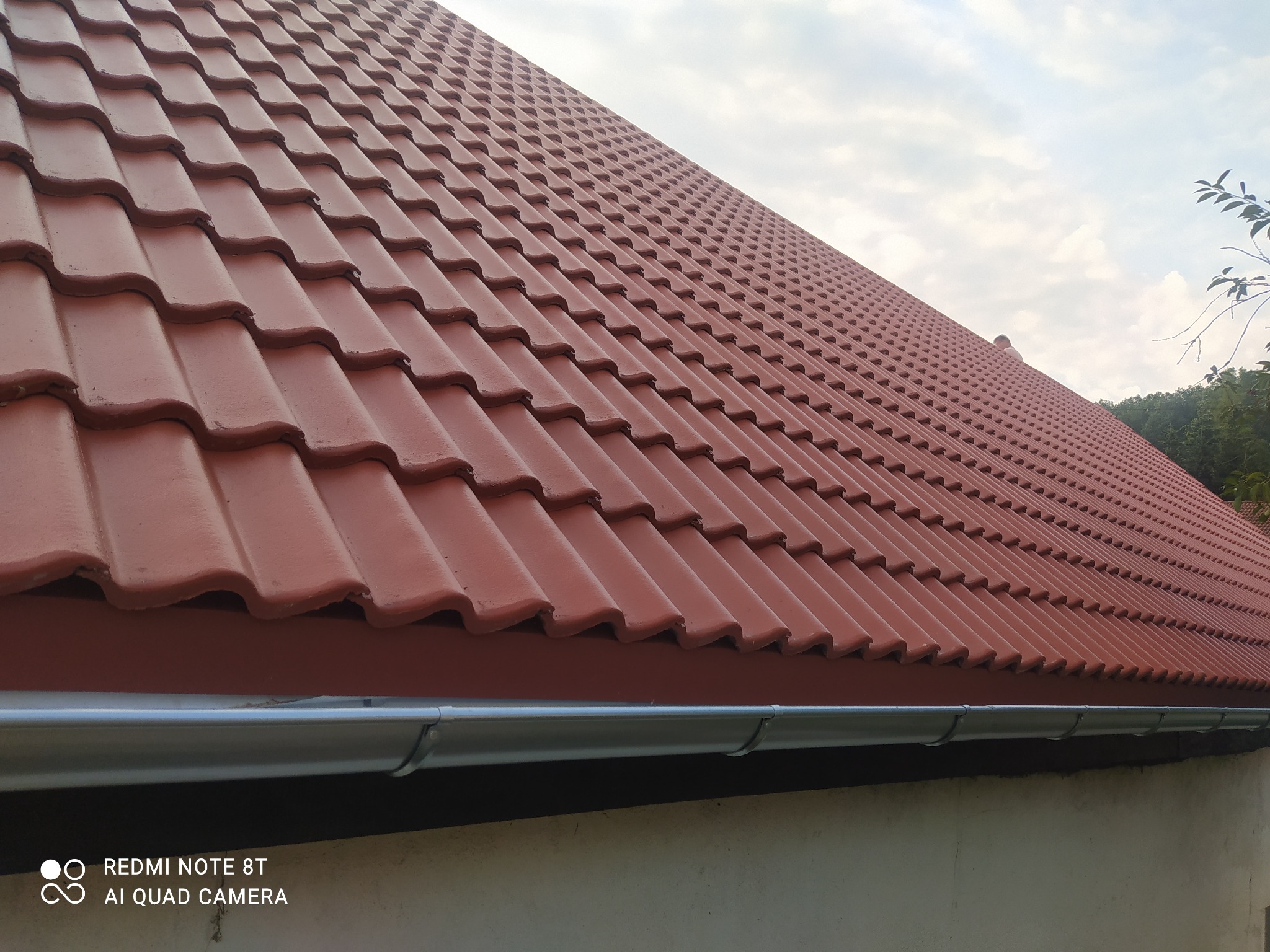 Czerwona dachówka ceramiczna na skośnym dachu, widoczne rynny z blachy ocynkowanej, fragment elewacji budynku i błękitne niebo z chmurami w tle.