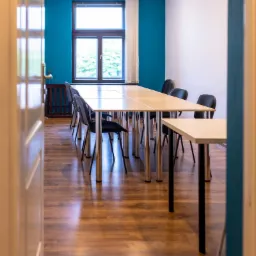 Jasna sala konferencyjna z długim stołem, krzesłami na chromowanych nogach, turkusową ścianą i widokiem z okna na zieleń, widziana zza uchylonych drzwi.