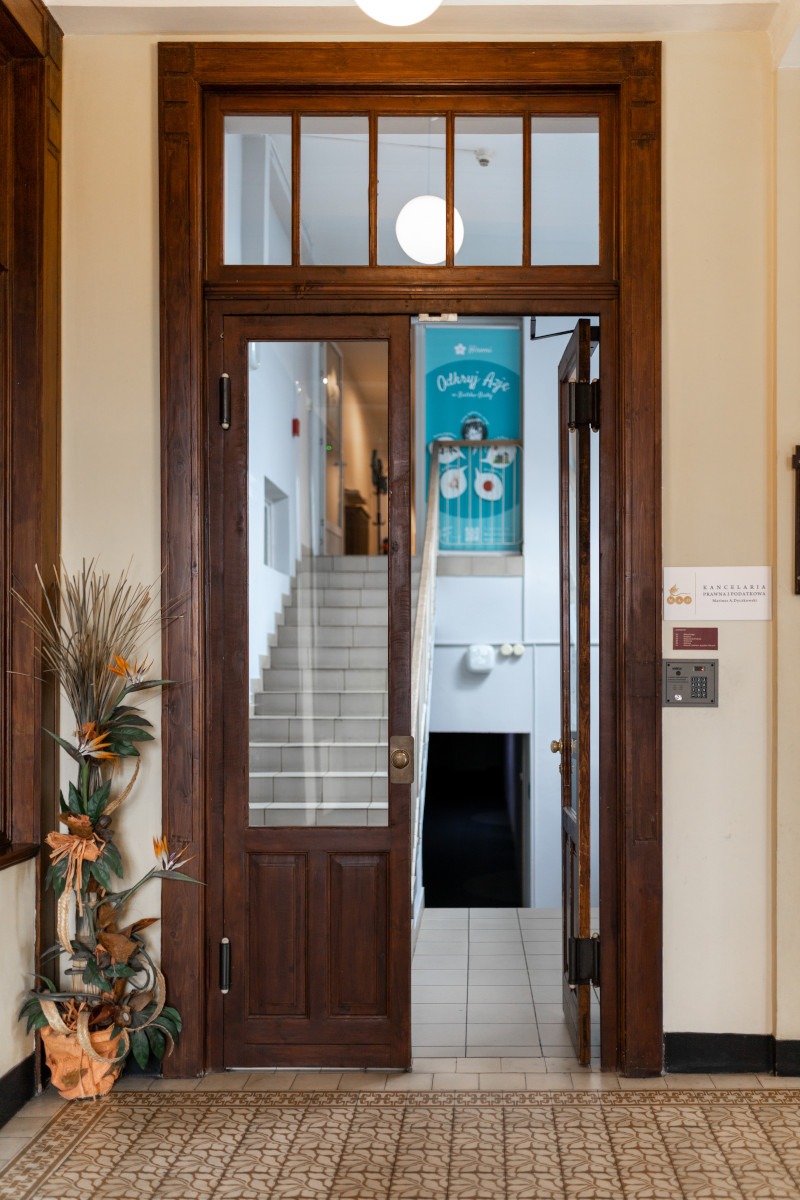 Wejście do budynku z drewnianymi drzwiami z szybami, widoczne schody, plakat reklamowy na ścianie i dekoracja kwiatowa w rogu.