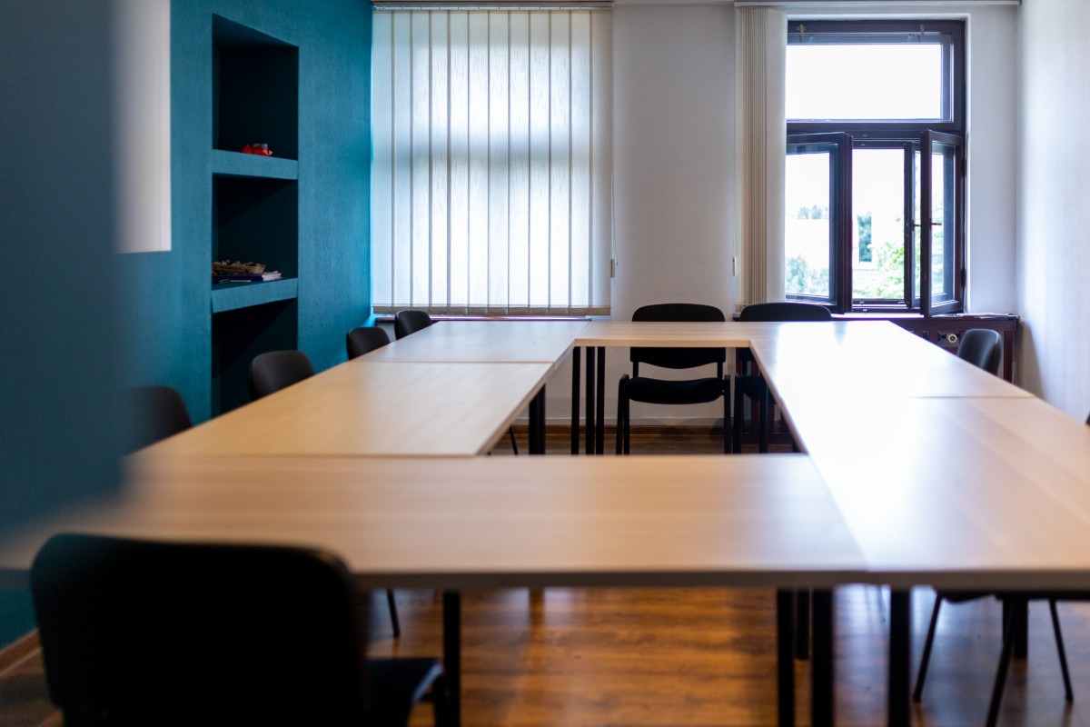 Sala lekcyjna z ustawionymi w kształt litery U jasnymi stołami i czarnymi krzesłami na drewnianej podłodze, turkusowa ściana z wnękami z lewej strony, okno z otwartymi skrzydłami w tle.