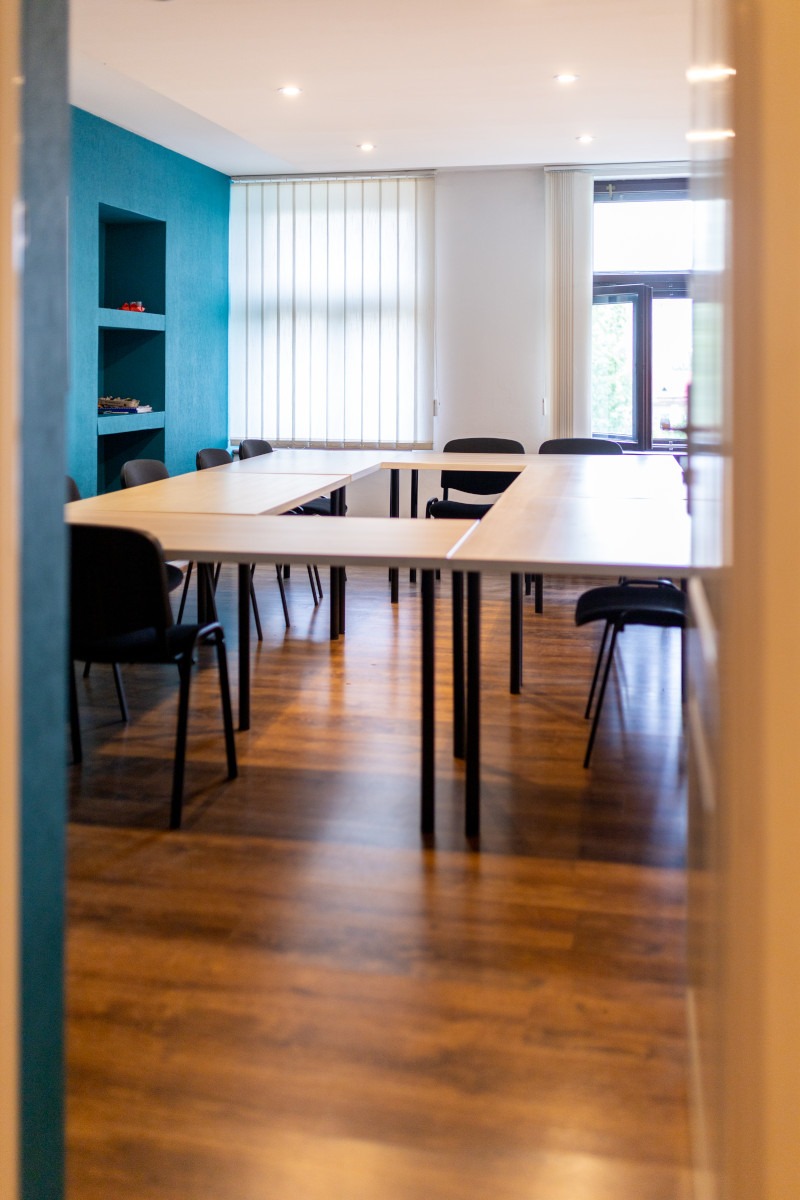 Sala konferencyjna z ustawionymi w podkowę stołami i czarnymi krzesłami, widok zza framugi drzwi, drewniana podłoga, niebieska ściana z wnękami w tle.