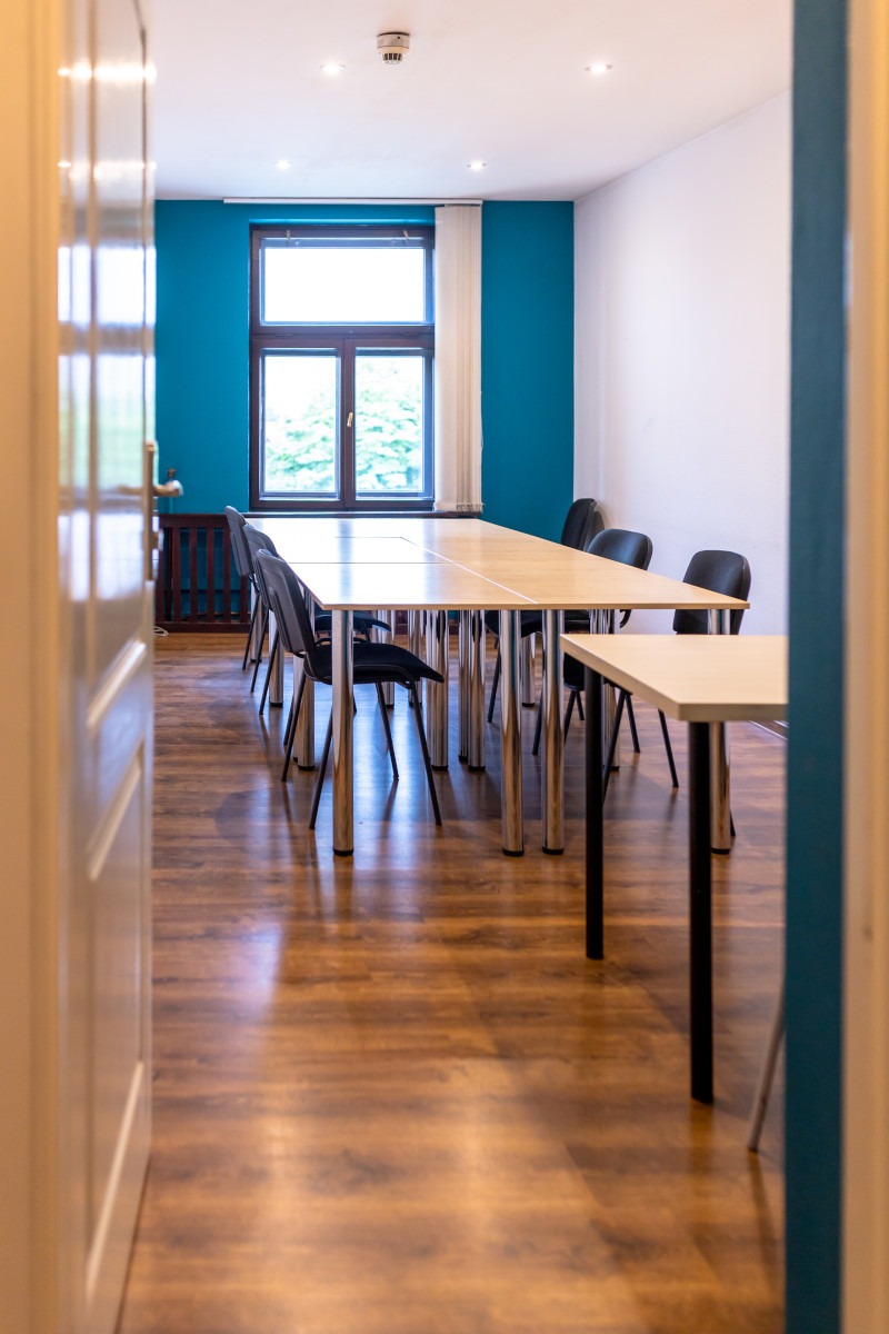 Jasna sala konferencyjna z długim stołem, krzesłami na chromowanych nogach, turkusową ścianą i widokiem z okna na zieleń, widziana zza uchylonych drzwi.