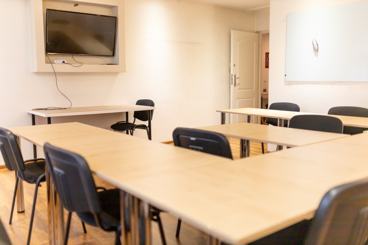 Jasna sala lekcyjna z drewnianymi biurkami i czarnymi krzesłami, tablica interaktywna na ścianie, neutralne kolory, przygotowana do zajęć.