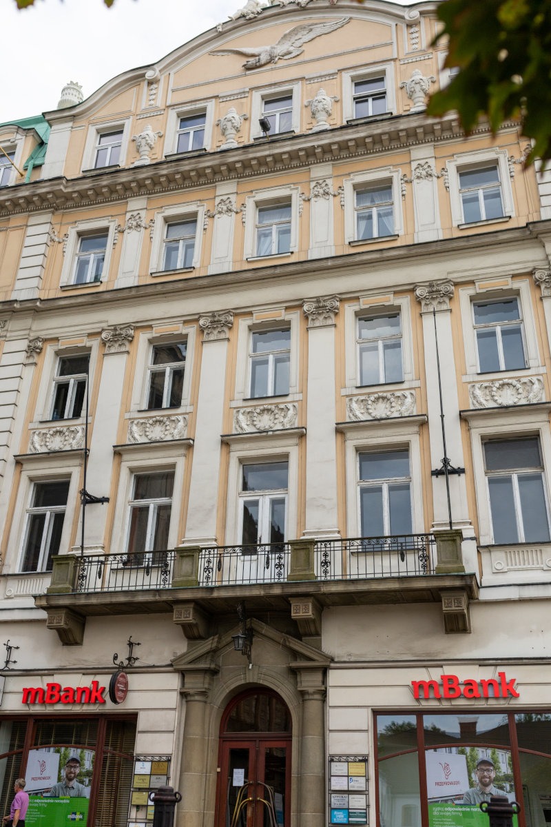 Fasada kamienicy w stylu klasycystycznym z balkonem, zdobieniami i rzeźbą orła na szczycie, poniżej widoczne wejście do budynku i szyldy mBank.