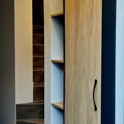 Wbudowana szafa z drewnianymi drzwiami przesuwnymi i otwartymi półkami, widoczne schody w tle.