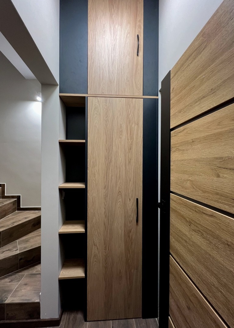 Wysoka zabudowa z drewna w odcieniach beżu i czerni, obejmująca szafę z długimi uchwytami, otwarte półki i drzwi z ukośnymi wstawkami, z widocznymi fragmentami schodów w tle.