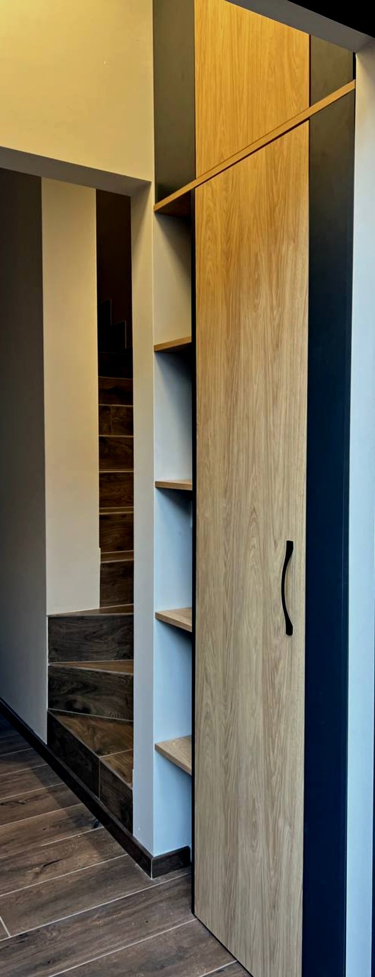 Wbudowana szafa z drewnianymi drzwiami przesuwnymi i otwartymi półkami, widoczne schody w tle.