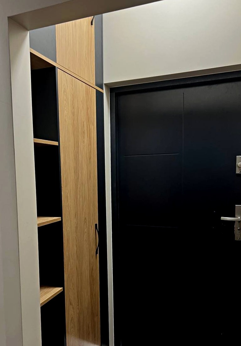Wysoka szafa z jasnego drewna z czarnymi akcentami, wbudowana obok czarnych drzwi wejściowych, widok z bliska na wykończone wnętrze.