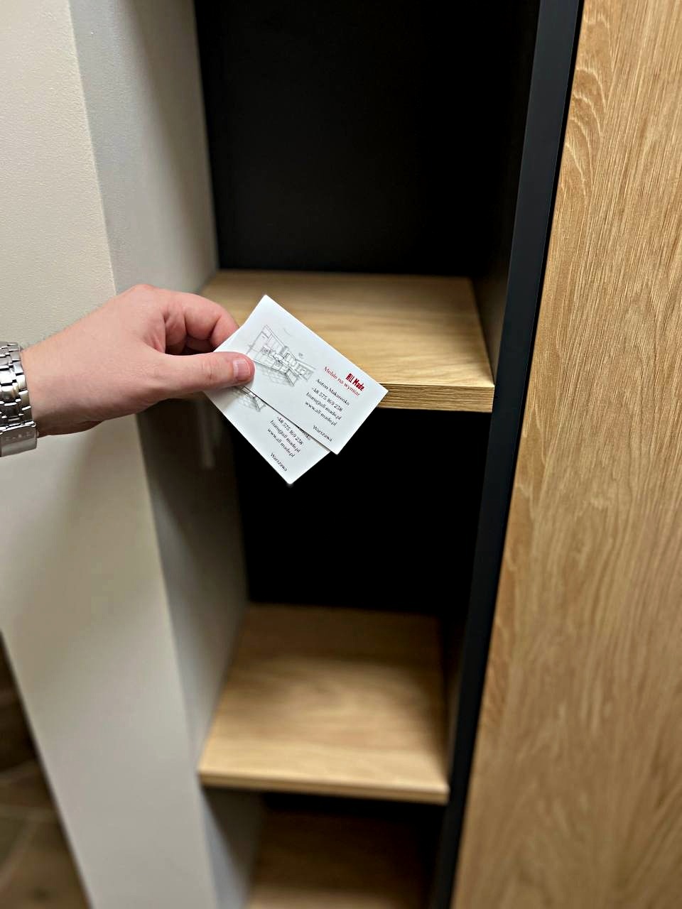 Ręka trzymająca wizytówkę firmy meblarskiej na tle półek w szafie z drewnianymi elementami i czarnymi obramowaniami.