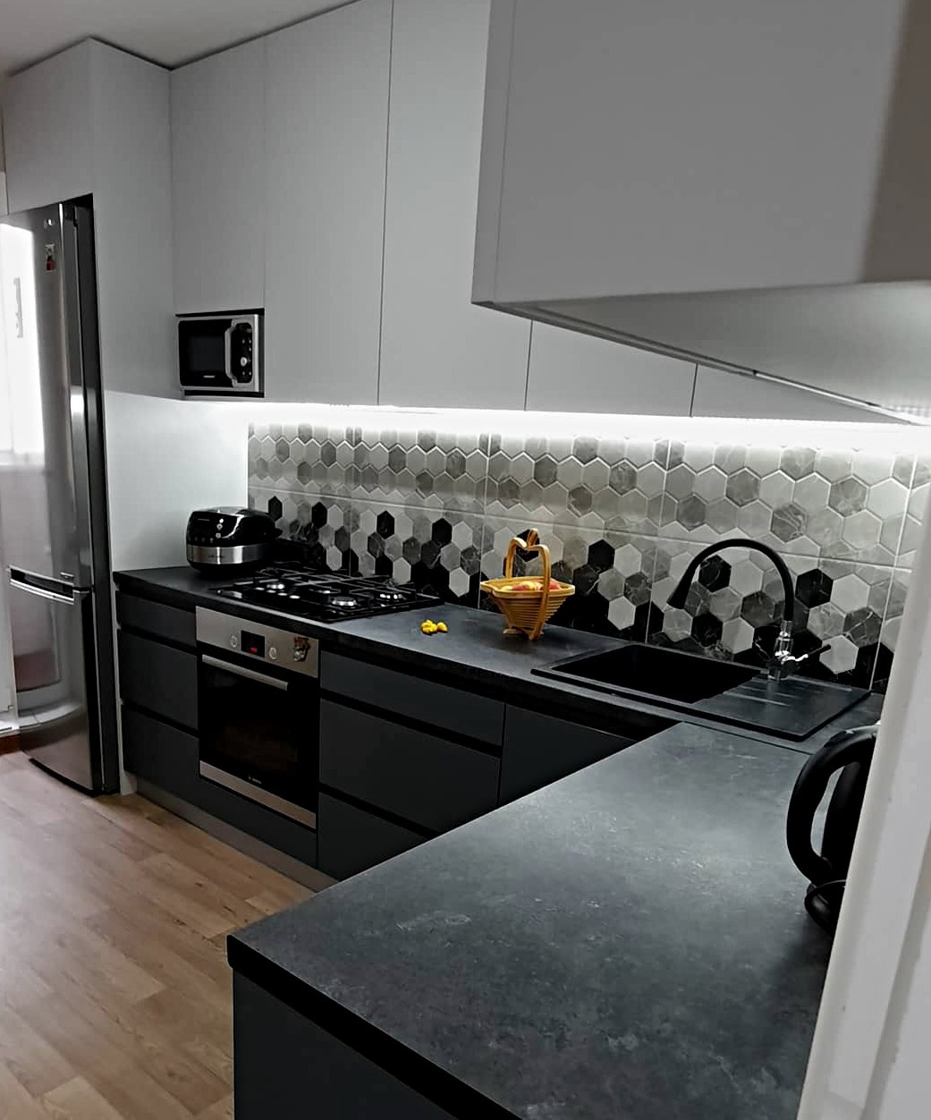 Nowoczesna kuchnia z szarymi szafkami, czarnym blatem i heksagonalnymi płytkami na ścianie, oświetlona listwą LED pod szafkami wiszącymi.