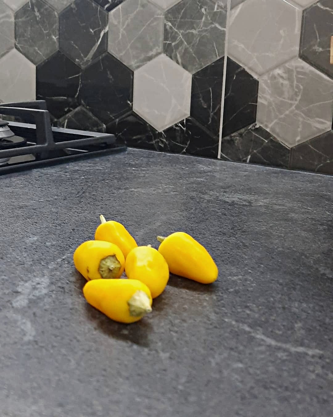Czarny blat kuchenny z marmurkowym wzorem i heksagonalnymi płytkami na ścianie, na którym leży kilka żółtych papryk.