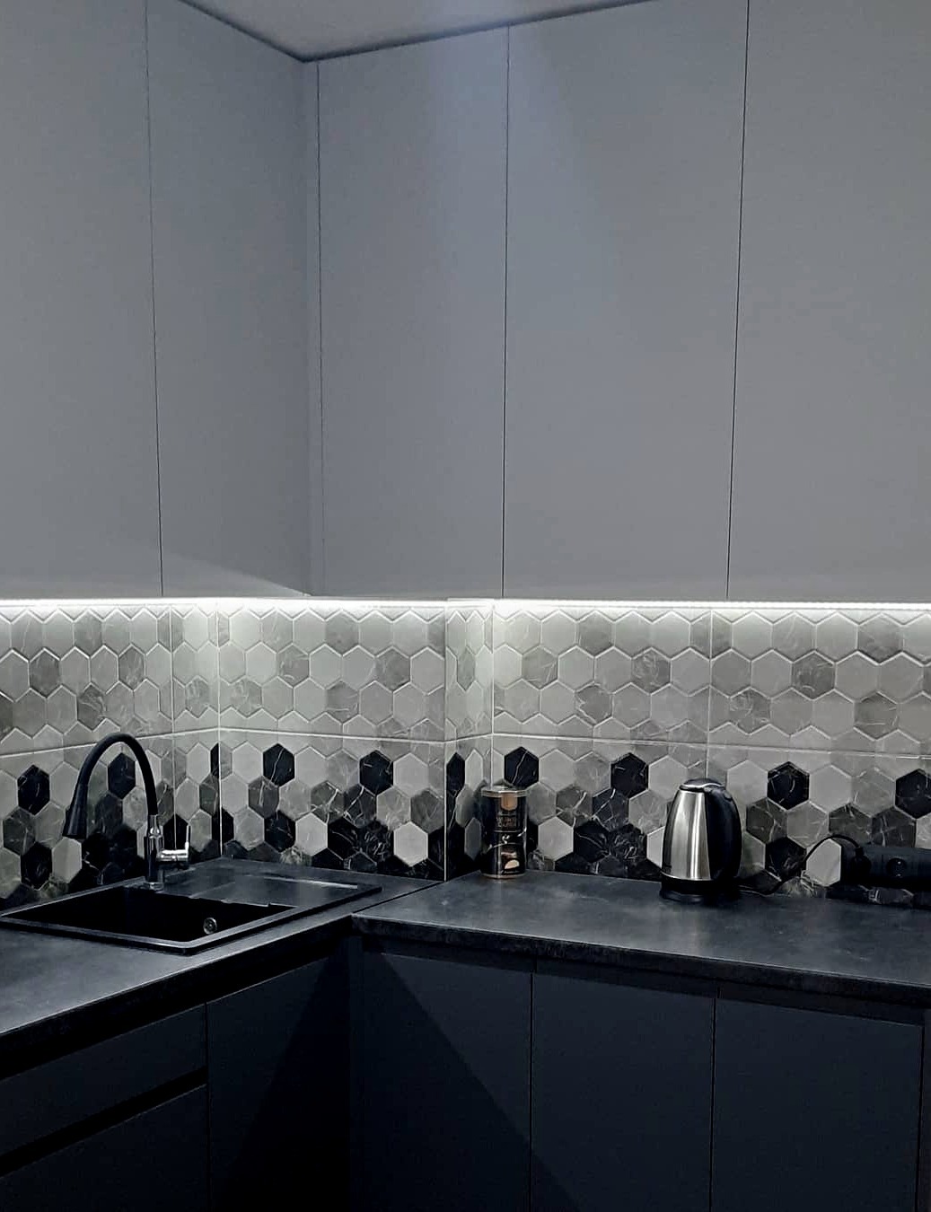 Nowoczesna kuchnia z szarymi szafkami, czarnym blatem i heksagonalnymi płytkami nad zlewem, podświetlona listwą LED.