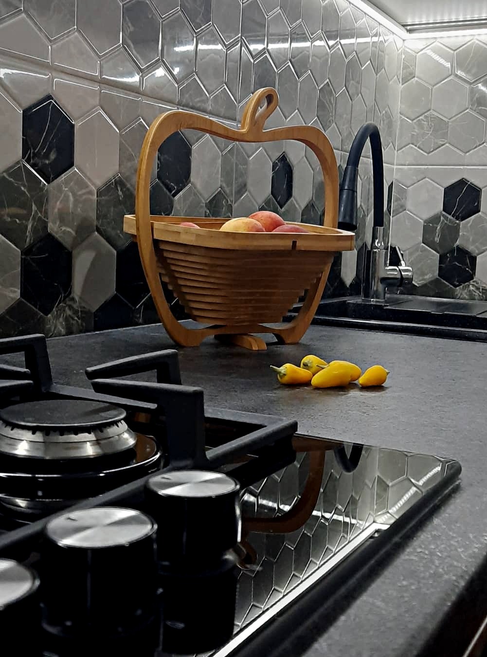 Nowoczesna kuchnia z czarnym blatem, płytą indukcyjną i drewnianym koszykiem w kształcie jabłka wypełnionym owocami, na tle mozaiki heksagonalnej w odcieniach szarości i czerni.