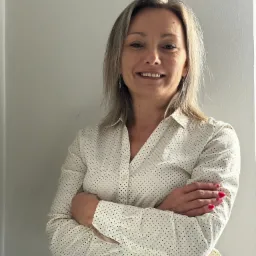 Uśmiechnięta kobieta w koszuli w kropki, z założonymi rękami, prezentuje profesjonalny wizerunek. Jasne tło, naturalne światło, widoczne detale makijażu i biżuterii.