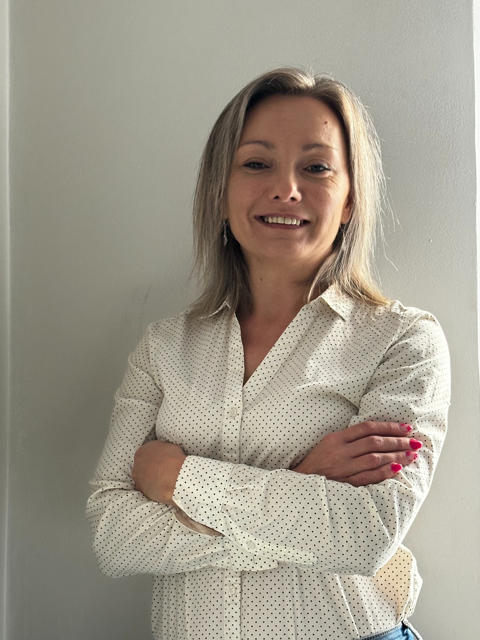 Uśmiechnięta kobieta w koszuli w kropki, z założonymi rękami, prezentuje profesjonalny wizerunek. Jasne tło, naturalne światło, widoczne detale makijażu i biżuterii.