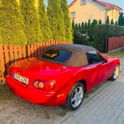 Czerwony kabriolet Mazda Miata z czarnym dachem zaparkowany na kostce brukowej przed domem z zielonymi tujami w tle.