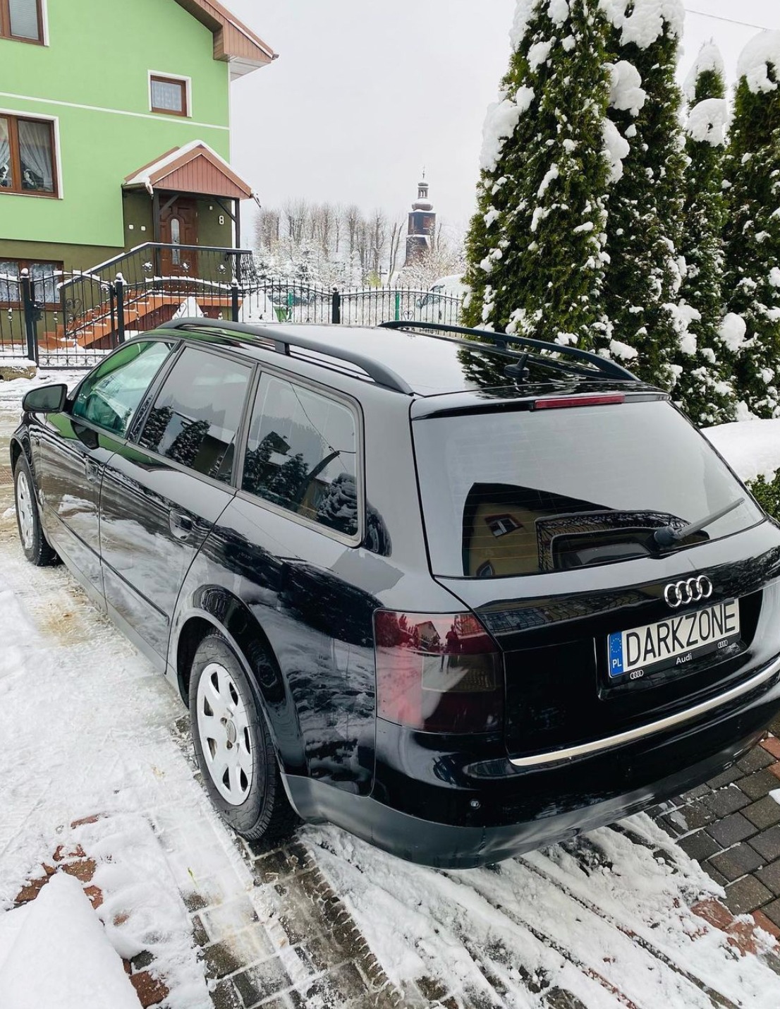 Czarne kombi Audi z przyciemnionymi szybami i lampami, zaparkowane na śniegu przed zielonym domem w zimowy dzień.