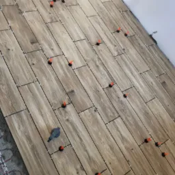 Układanie podłogi z płytek imitujących drewno z użyciem systemu poziomowania, widoczne krzyżyki dystansowe i pomarańczowe kliny, w tle fragment ściany i ogrzewanie podłogowe.