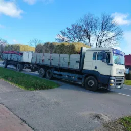 Ciężarówka MAN z przyczepą transportuje bele siana po asfaltowej drodze w słoneczny dzień.