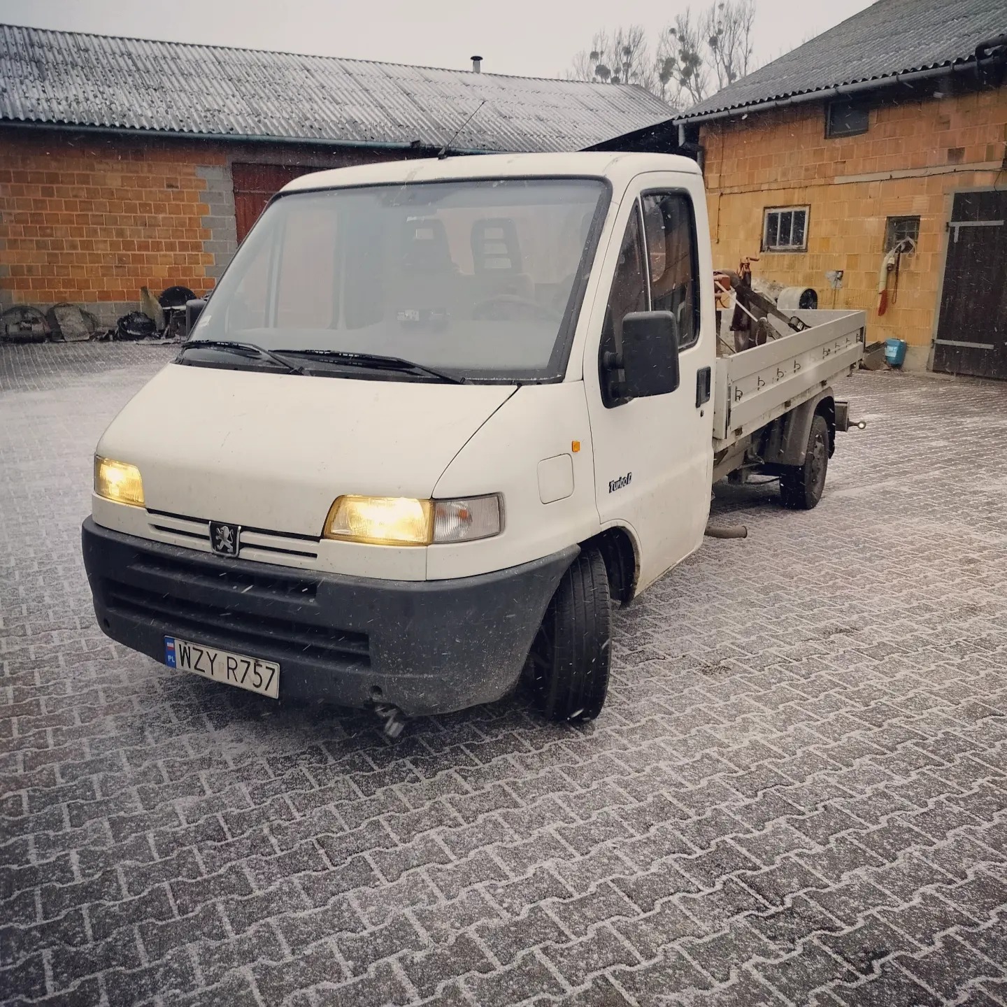 Biały samochód dostawczy Peugeot TurboD z otwartą skrzynią ładunkową, zaparkowany na kostce brukowej w zimowy dzień. Na skrzyni ładunkowej widoczne narzędzia i akcesoria.