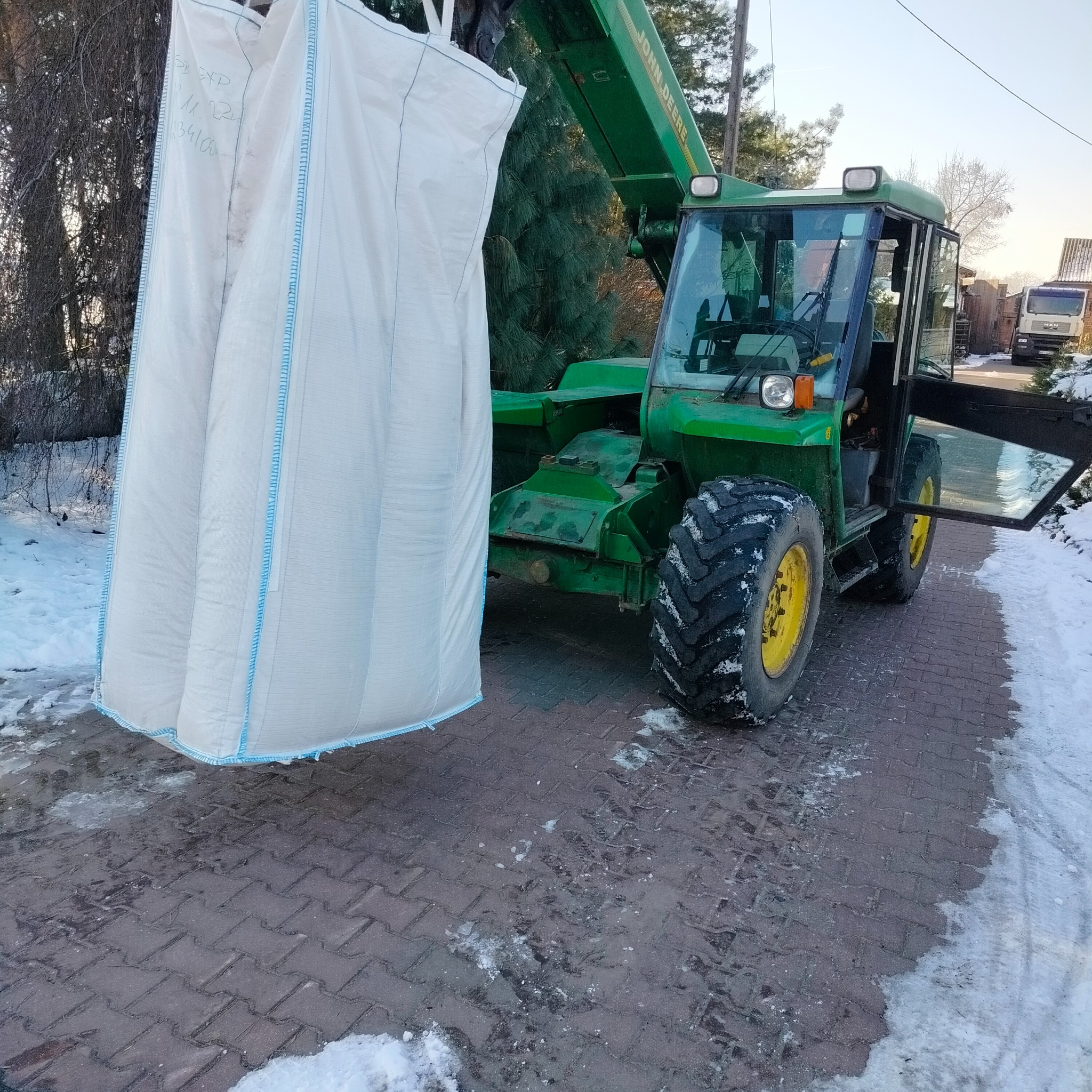 Zielony wózek widłowy John Deere podnosi dwa duże białe worki z niebieskimi szwami na tle ośnieżonej kostki brukowej i drzew
