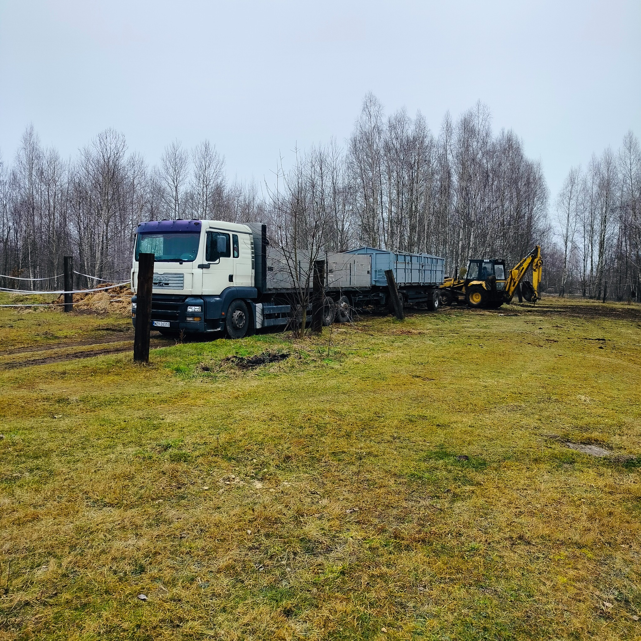 Ciężarówka MAN z przyczepą i żółta koparka na trawiastym terenie z drzewami w tle, widoczne ślady opon i słupki ogrodzeniowe.