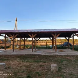 Drewniana wiata z czerwonym dachem z blachy trapezowej, wsparta na słupach, na betonowej wylewce, w tle pole i drzewa, widoczne elementy konstrukcyjne dachu.