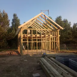 Wznoszenie drewnianej konstrukcji dachu budynku mieszkalnego w otoczeniu drzew, widoczne przygotowane deski i pracownik na rusztowaniu.