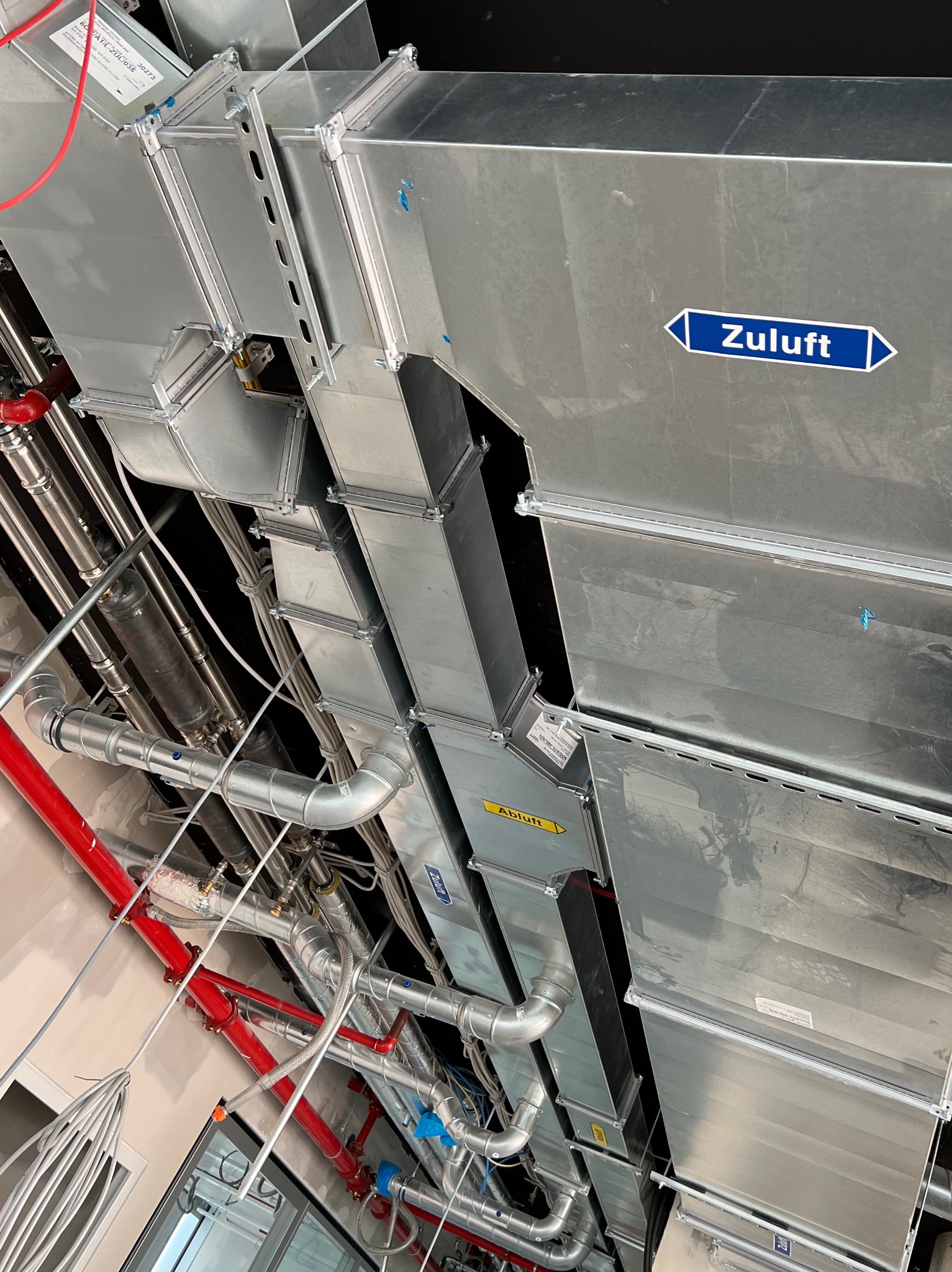 Widok z dołu na zainstalowany system kanałów wentylacyjnych z etykietami 'Zuluft' i 'Abluft', oraz rury instalacji przeciwpożarowej w kolorze czerwonym.