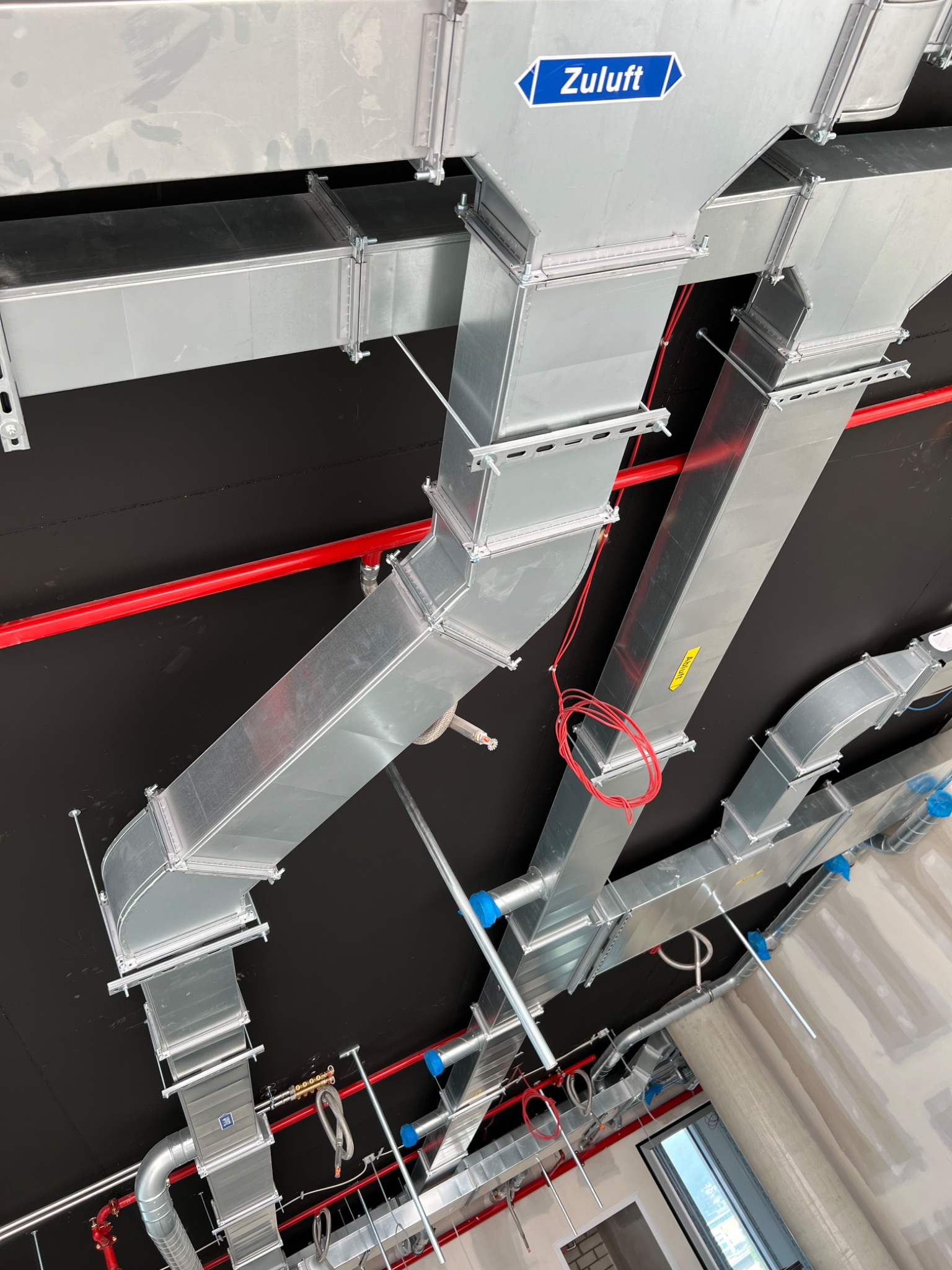 Srebrne kanały wentylacyjne pod czarnym sufitem z widocznymi czerwonymi rurami, niebieskimi zaślepkami i etykietą 'Zuluft'.