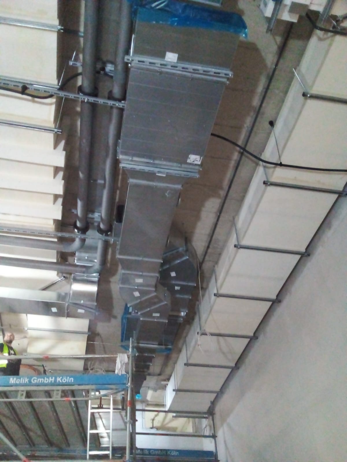 Widok z dołu na montaż systemu kanałów wentylacyjnych z blachy ocynkowanej, prowadzonych pod sufitem budynku w stanie surowym, widoczne elementy rusztowania i instalacji elektrycznej.