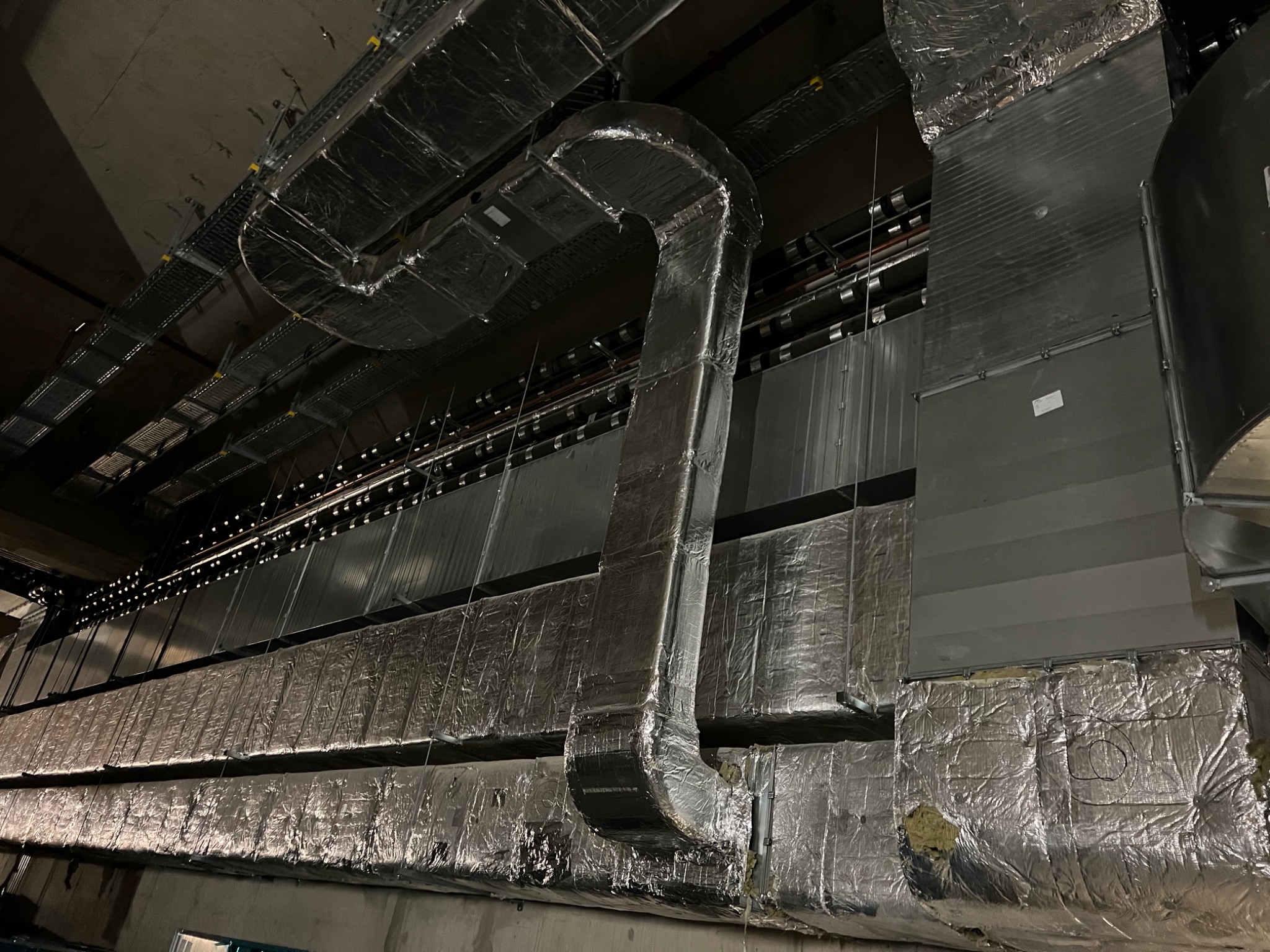 Widok na system kanałów wentylacyjnych z izolacją aluminiową, zamontowanych pod sufitem w przestrzeni przemysłowej; widoczne połączenia i mocowania.