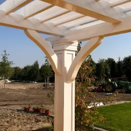 Fragment białej drewnianej konstrukcji altany z widocznym słupem i belkami, na tle przygotowanego terenu z traktorem i zielenią, zabezpieczona folią ochronną przed zabrudzeniem.