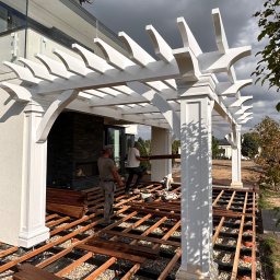 Usługi Ogólnobudowlane - Montaż drewnianego tarasu z białą pergolą przy nowoczesnym domu; widoczny szkielet konstrukcji, deski tarasowe, narzędzia i dwóch pracowników w trakcie pracy.