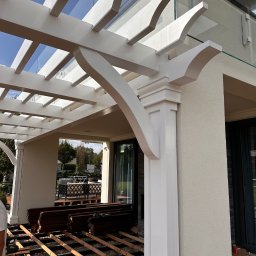 Usługi Ogólnobudowlane - Biała pergola z elementami szklanymi, widok na konstrukcję tarasu w trakcie budowy, deski ułożone na legarach, narzędzia pracy w tle, jasne otoczenie domu.