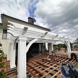 Usługi Ogólnobudowlane - Budowa tarasu drewnianego z białymi kolumnami pod zadaszeniem, widoczne legary, deski, piła tarczowa, pracownicy w trakcie montażu, nowoczesny dom w tle, pochmurne niebo.