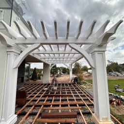 Usługi Ogólnobudowlane - Budowa tarasu: drewniana konstrukcja pod taras z białymi kolumnami i fragmentami desek, widoczne narzędzia i pracownicy w tle, kamieniste podłoże.