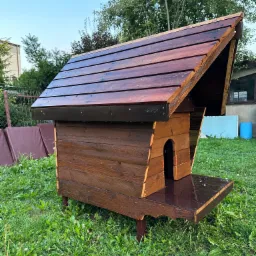 Nowa, brązowa buda dla psa z drewnianym daszkiem, stojąca na trawie w ogrodzie, po deszczu.