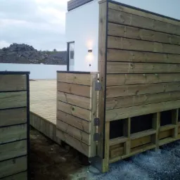 Nowoczesny taras z drewnianą zabudową, widoczne elementy konstrukcyjne i fragment budynku z oknem, w tle usypisko kamieni.