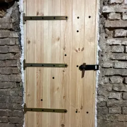 Nowe drewniane drzwi z metalowymi zawiasami i czarnym zamkiem, osadzone w starej, ceglanej ścianie z widoczną pianką montażową uszczelniającą szczelinę.