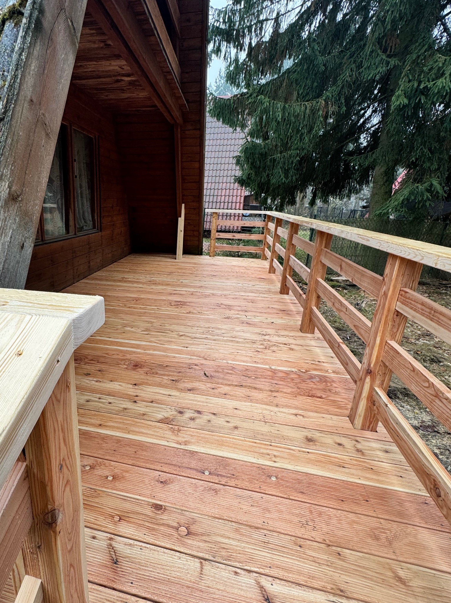 Nowy, drewniany taras przylegający do drewnianego domu, z widoczną balustradą i schodami, w tle drzewa iglaste i dach sąsiedniego budynku.