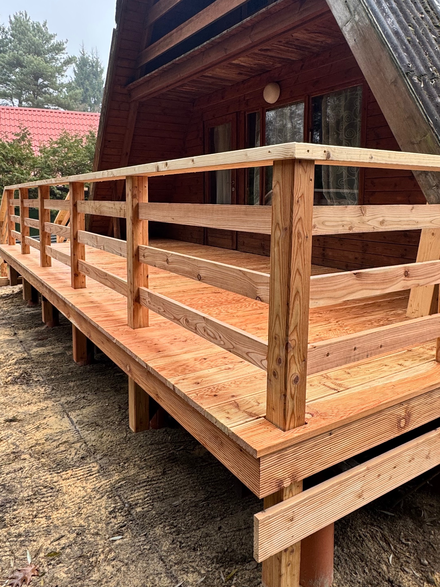 Nowo wybudowany drewniany taras z balustradą przy domku w stylu A-frame, surowe drewno z widocznym usłojeniem, pomalowane na jasny odcień, widoczne podpory tarasu na fundamencie.