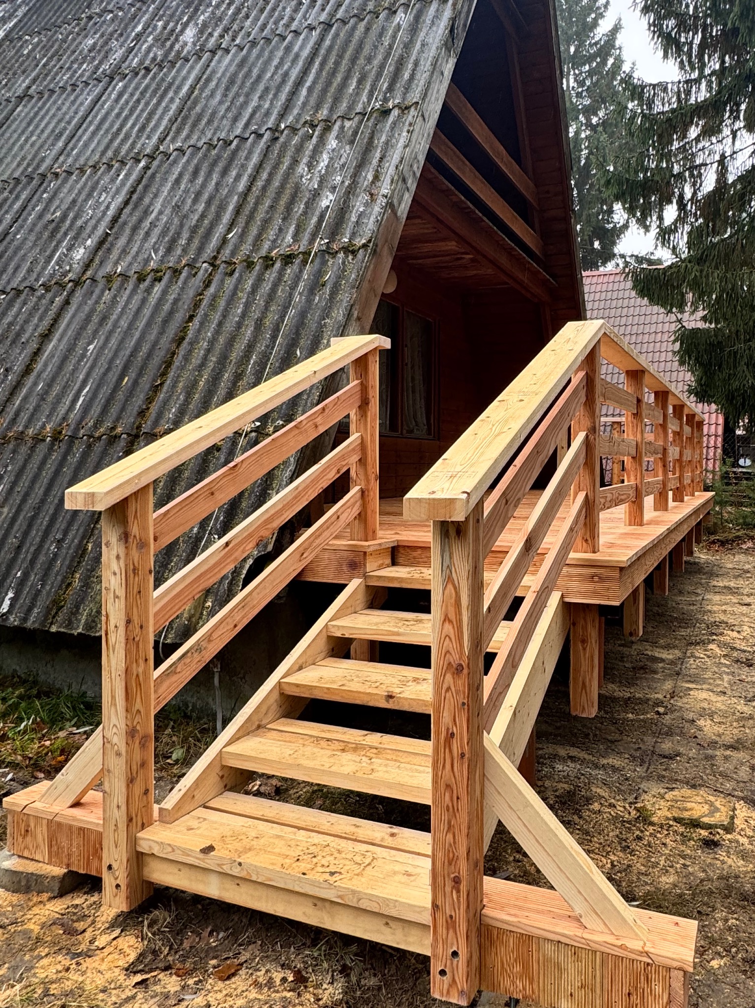 Nowe, surowe drewniane schody i taras przed drewnianym domkiem letniskowym z szarym, falistym dachem, widok z dołu pod kątem.