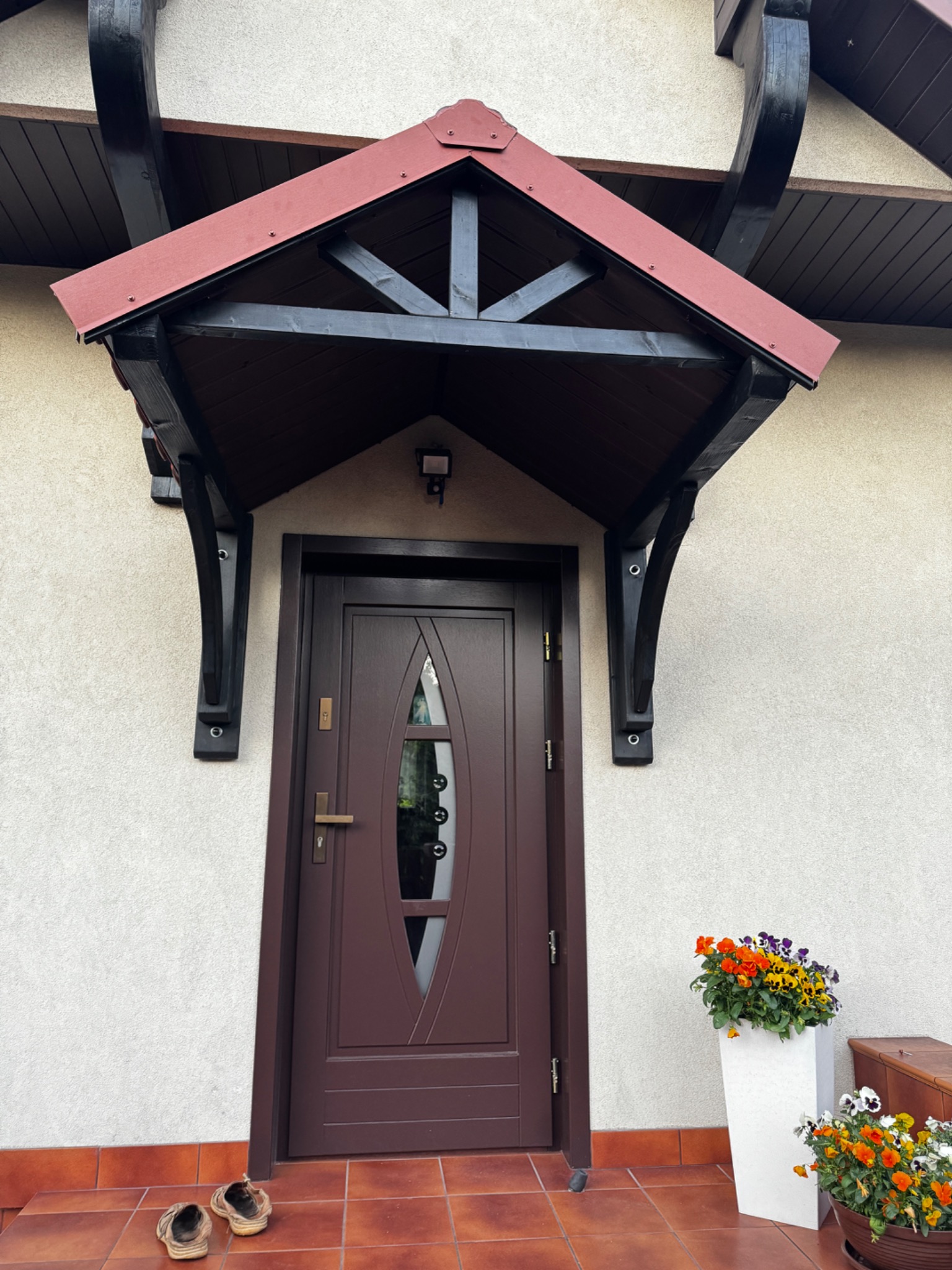 Brązowe drzwi wejściowe z daszkiem i ozdobnymi wspornikami. Na wycieraczce leżą klapki, obok donice z kwiatami. Elewacja budynku w jasnym odcieniu.