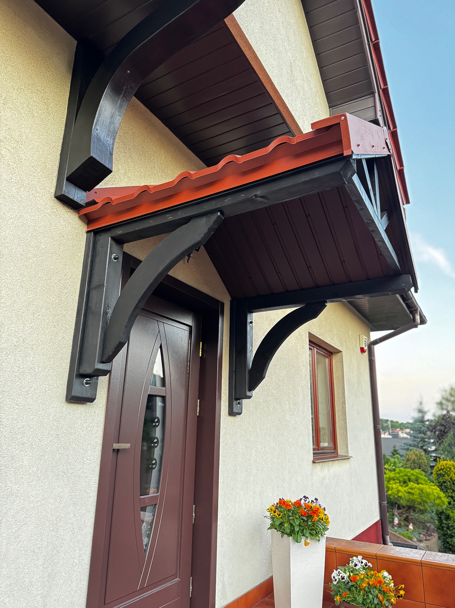 Zadaszenie nad brązowymi drzwiami wejściowymi, wsparte na czarnych, drewnianych wspornikach. Czerwona dachówka, kremowa elewacja budynku, kwiaty w donicach.