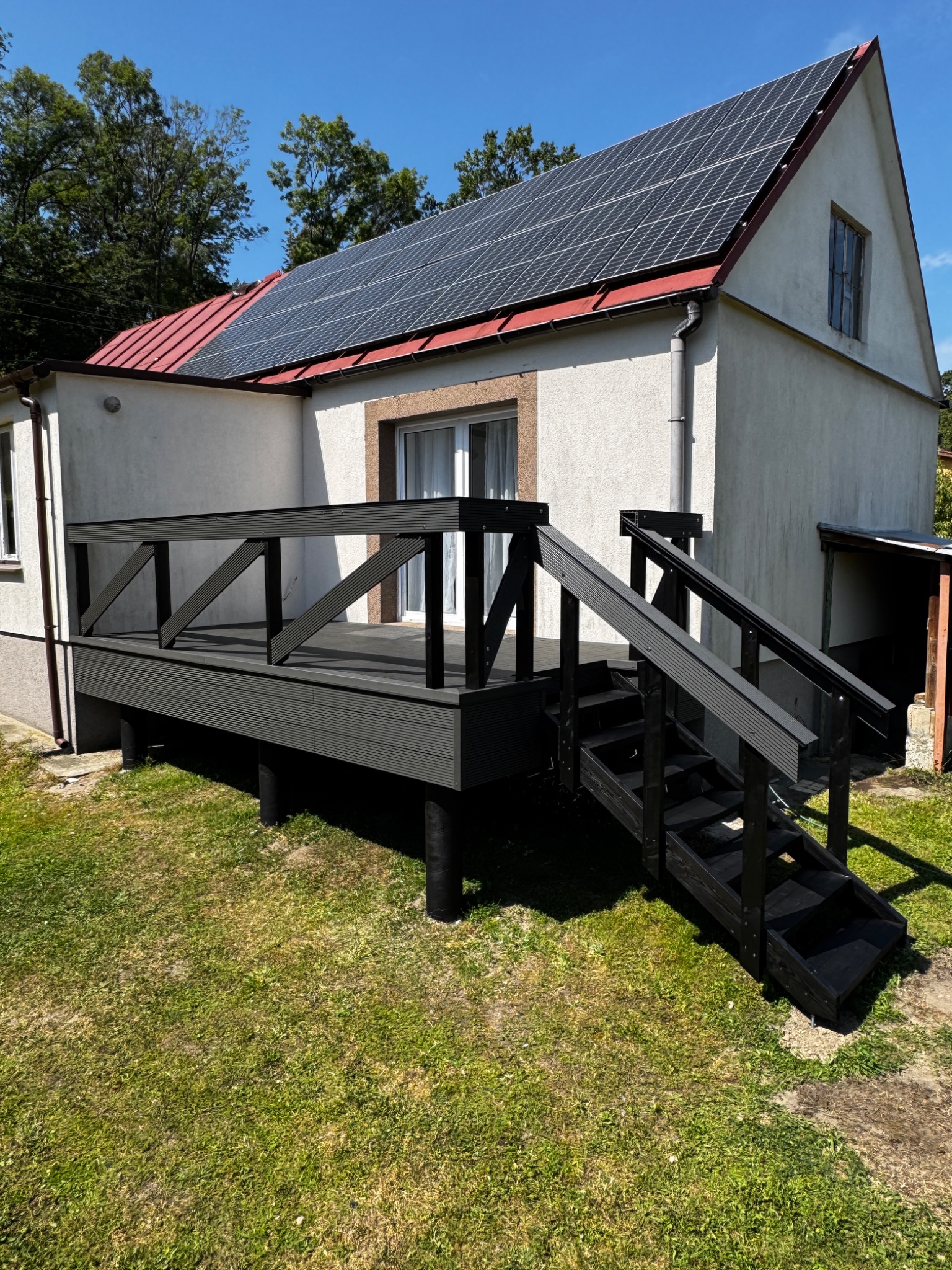 Nowoczesny taras kompozytowy w kolorze antracytowym z schodami i balustradą, przylegający do domu z panelami słonecznymi na dachu. Słoneczny dzień, zielony trawnik.