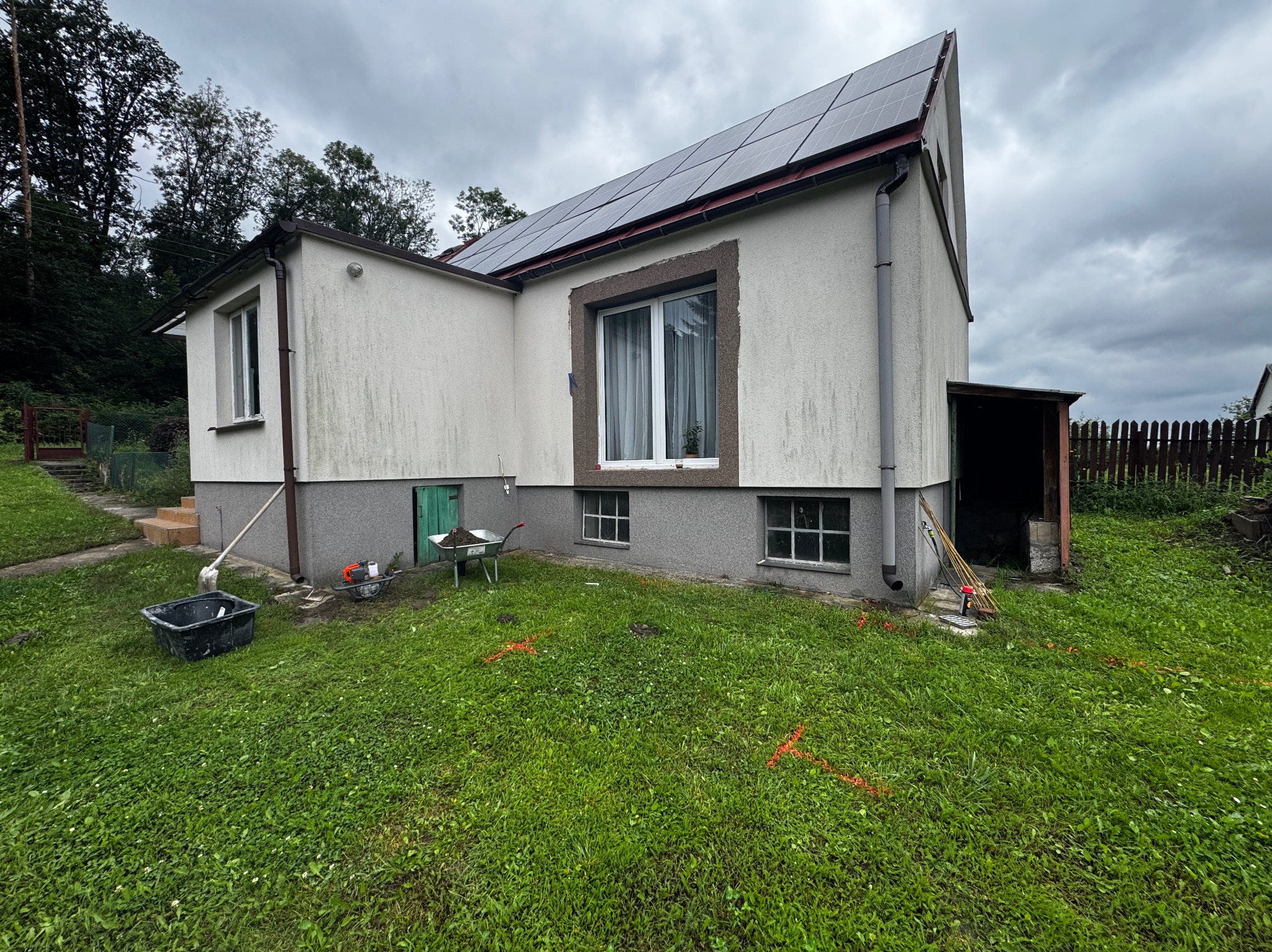 Dom z panelami słonecznymi, widoczne narzędzia ogrodowe, sugerujące prace ziemne lub przygotowanie terenu. Pochmurne niebo, trawa z pomarańczowymi oznaczeniami.