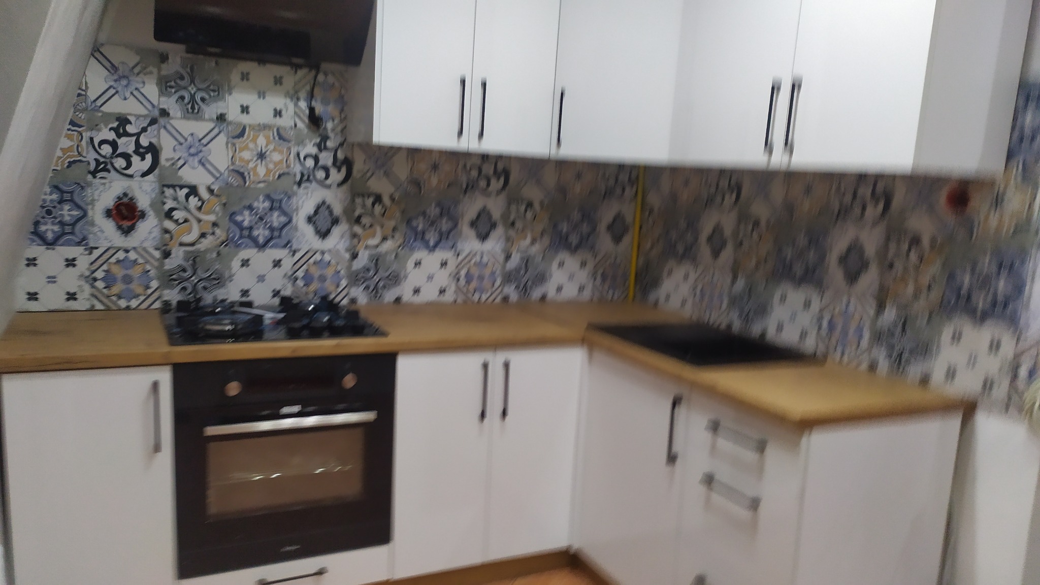 Fragment białej kuchni z drewnianym blatem, ciemnymi uchwytami i ozdobnymi płytkami na ścianie, widoczny piekarnik i płyta gazowa.