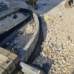 Budowa obrzeża rabaty z szarej kostki granitowej na podbudowie z grysu i zaprawy, widoczna taczka i mini koparka w tle.